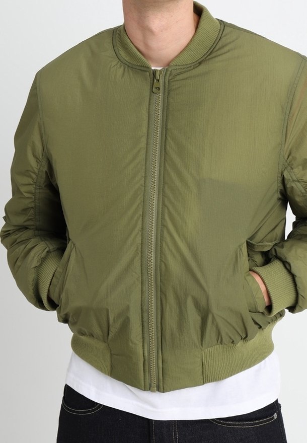 Homme portant une veste bomber vert olive à fermeture éclair, avec col, poignets et bas côtelés, mains dans les poches, par-dessus une chemise blanche et un pantalon foncé.