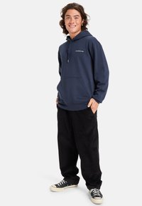 Sudadera con capucha de color azul marino, con un bolsillo tipo canguro, cordón en la capucha y detalle de logotipo. Combinada con pantalones de pana negros y zapatillas negras.