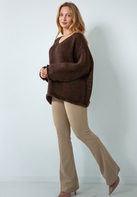 Brązowy, oversize'owy sweter dzianinowy z dekoltem w kształcie V i ribbową teksturą, zestawiony z beżowymi dopasowanymi spodniami i jasnobrązowymi szpilkami.