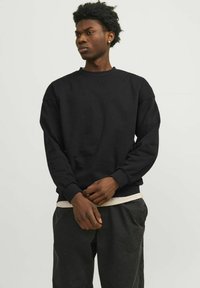 Must olnud puuvillane must crewneck kootud kampsun. Omab soonikute krae ja alumist osa, esiküljel on tikkimine ning lahtine istuvus. Kantud hallide pükste peal.