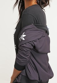 Maglietta nera a maniche lunghe indossata sotto una giacca sportiva viola con zip. La giacca presenta un motivo testurizzato e un logo bianco sulla manica.