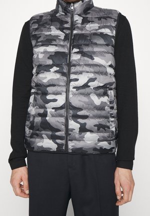 Homme portant un gilet matelassé camouflage gris et noir par-dessus une chemise noire à manches longues avec un pantalon noir.