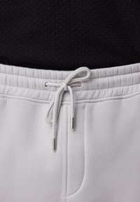 Gros plan sur la ceinture d'un pantalon de survêtement gris clair avec un cordon noué et des embouts métalliques, porté avec un haut noir texturé.
