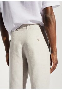 Pantaloni in lino beige chiaro con vestibilità dritta, caratterizzati da due tasche posteriori e un dettaglio in bottone, abbinati a una camicia bianca a maniche corte.