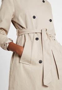 Cappotto beige con design doppiopetto, bottoni neri, cintura abbinata e tasche a toppa. Presenta dettagli a motivo sui polsini.