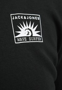 Crna majica kratkih rukava s bijelim grafičkim logotipom na kojem piše "JACK&JONES" i "WAVE SURFER", prikazuje surfera i stilizirane sunčeve zrake.