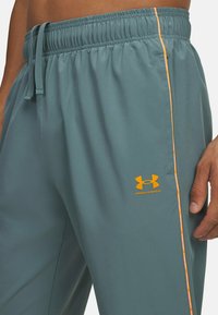 Grå atletiske bukser med elastisk talje, prydet med en orange lodret stribe og et Under Armour-logo i orange på venstre ben.