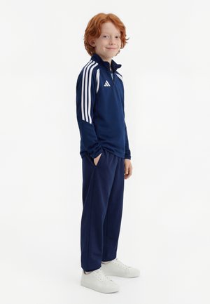 Punapea noormees seisab käed taskutes, seljas tumesinine Adidas dressipluus ja valged tossud, valgel taustal.