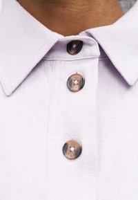 Φωτεινό μωβ πουκάμισο με γιακά και τρία ορατά μαρμάρινα καφέ κουμπιά κουμπωμένα μπροστά.