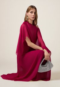 Dieprode avondjurk met doorschijnende cape-mouwen, zachte textuur en een vloeiende silhouet. Gecombineerd met een kleine handtas met zilveren kralen.