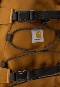 Καφέ υφασμάτινο σακίδιο με μαύρα λουριά και μεταλλικά μέρη. Σημαντικές λεπτομέρειες περιλαμβάνουν ένα γκρι ελαστικό κορδόνι και μια λευκή ετικέτα με λογότυπο.