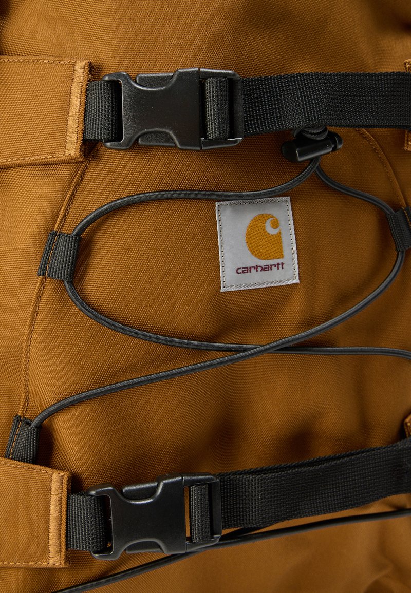 Sac à dos en tissu marron avec des bretelles et des accessoires noirs. Détail notable : un cordon élastique gris et une étiquette blanche avec un logo.