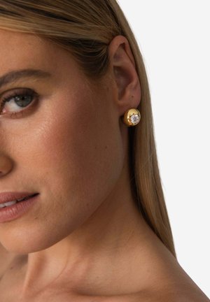 Runder Gold-Stecker mit einem klaren, großen Edelstein in der Mitte, vor einem neutralen Hautton-Hintergrund. Glatte, reflektierende Oberfläche.