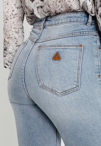 Ljust blå denimjeans med en liten brun triangel-logotyp på bakfickan, som bärs med en mönstrad långärmad skjorta instoppad.