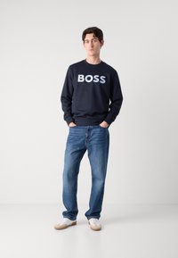 Nāvzila džemperis ar "BOSS" logotipu gaiši pelēkā krāsā, kombinēts ar zilu džinsu biksēm un baltām kedām. Gluda auduma un brīva piegriezuma.