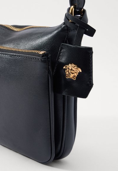 Borsa a tracolla in pelle nera con zip dorata, design elegante e un emblema di leone attaccato sul fronte. Texture liscia e forma strutturata.