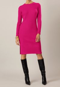 Abito a maglia a coste di un vivace rosa, con maniche lunghe, scollo rotondo e uno spacco laterale, abbinato a stivali neri fino al ginocchio.