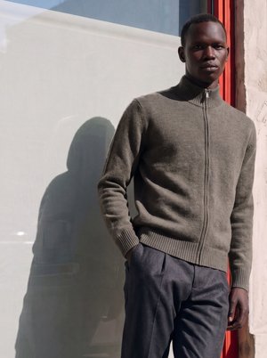 Jeune homme portant un pull marron zippé et un pantalon gris, debout avec une main dans la poche devant une paroi en verre montrant son ombre.