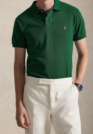 Mand iført mørkegrøn polo, beige bukser og armbåndsur med blå urskive og brun læderrem, hånd i lommen.