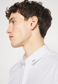 White collared shirt featuring a branded collar tag. Classic design with button closures, smooth texture, and a tailored fit.