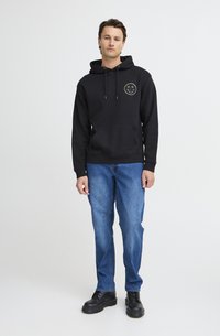 Felpa nera con tasca a marsupio frontale, cappuccio con coulisse regolabile e grafica circolare verde. Abbinata a jeans blu e scarpe nere.
