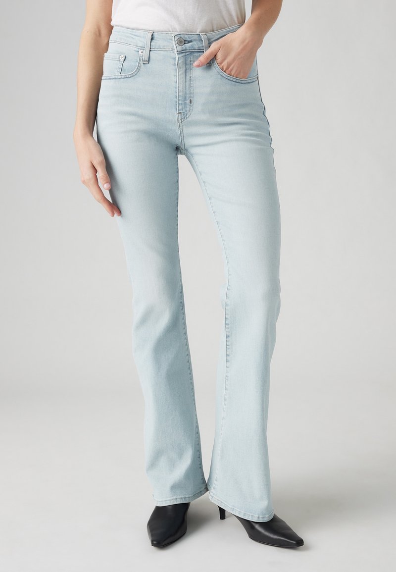 Femme portant un jean flare taille haute bleu clair avec poches avant, associé à des bottines noires à bout pointu.