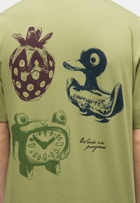 Πίσω μέρος ελιάς πράσινου πουκάμισου με εικονογραφήσεις: κόκκινη φράουλα, μπλε λαστιχένια πάπια με την επιγραφή «carhartt», πράσινο ρετρό ξυπνητήρι και το κείμενο «Work in progress».