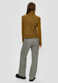 Braun gestrickter ärmelloser Pullover mit hohem Kragen, kombiniert mit grauen karierten weit geschnittenen Hosen und schwarzen flachen Schuhen. Rückansicht.