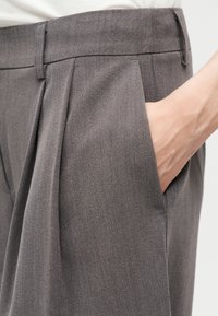 Main reposant dans la poche droite d'un pantalon gris plissé avec passants pour ceinture, visible de la taille au milieu de la cuisse.