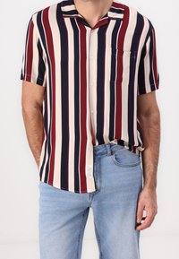 Adult male wearing a short-sleeve button-up shirt with vertical navy, beige, and burgundy stripes, paired with light blue jeans.