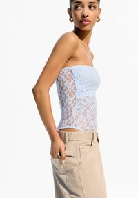 Svetlo modra top brez naramnic iz čipke z teksturiranim cvetličnim vzorcem, prilegajoča se oblika in gubana stranska podrobnost, v kombinaciji s bež hlačami.