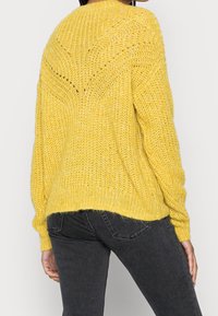 Eine Person trägt einen lockeren, gelben Strickpullover mit einem strukturierten Muster und schwarzen, figurbetonten Jeans, von hinten gezeigt.