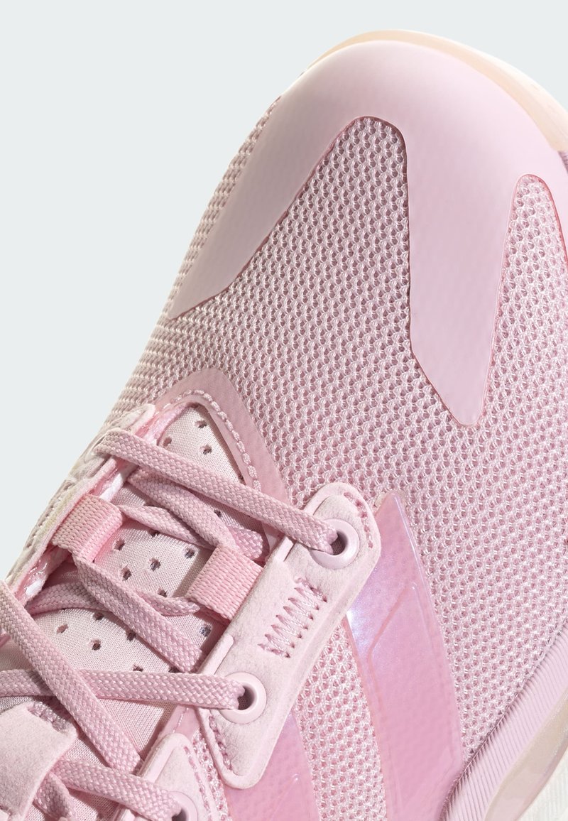 Zapatilla deportiva de color rosa claro con parte superior de malla, detalles de plástico y diseño con cordones. Presenta patrones texturizados y una puntera lisa.
