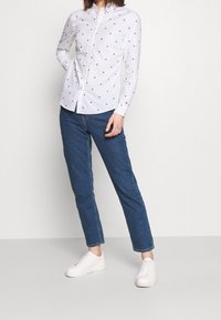 Vit skjorta med knappar och små röda och blå mönster, i kombination med blå jeans och vita sneakers. Tyget ser slätt och lätt ut.