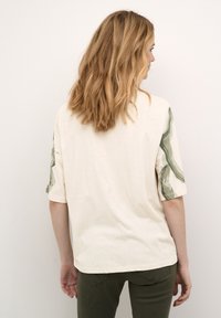 Camiseta oversized beige con mangas de diseño verde. Textura suave, cuello redondo y ajuste relajado. La vista trasera muestra tela lisa y dobladillo.