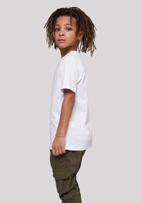 White cotton t-shirt with short sleeves, paired with olive green cargo pants. Child with dreadlocks stands in profile against a neutral background.