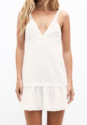 Biała sukienka z głębokim dekoltem w kształcie litery V, koronkowymi zdobieniami i regulowanymi ramiączkami. Posiada falbaniaste wykończenie na dole oraz lekką tkaninę.