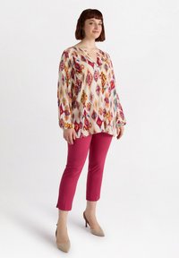 Mönstrad blus med lång ärm och V-ringning i krämfärg, prydd med livliga, flerfärgade geometriska mönster, kombinerad med rosa åtsittande byxor och beige klackskor.