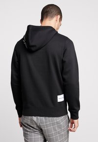 Černá mikina s kapucí, s bílým prošitím na výstřihu a bílou štítkem Calvin Klein na spodní pravé straně.