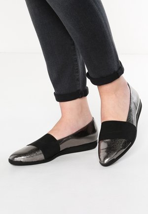 Pieds chaussés de ballerines métalliques à bout pointu avec des bandes élastiques noires, associées à un jean foncé retroussé, sur un sol blanc uni.