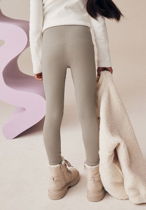 Geribbelde, lichtbruine legging met hoge taille, gecombineerd met beige winterlaarzen en een fluffy crèmekleurige jas. De achtergrond heeft zachte, gebogen vormen.