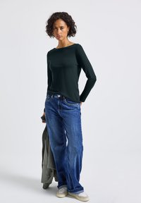 Donkergroene lange mouwen top in combinatie met blauwe wijde jeans. Draagt een grijze jas en witte sneakers. Eenvoudig ontwerp en relaxte pasvorm.