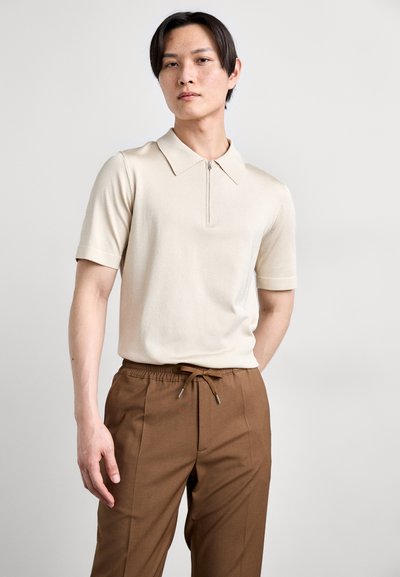 Lys beige kortærmet polo skjorte med en lynlås krave, parret med brune bukser med snøre og sidelommer samt en glat tekstur.