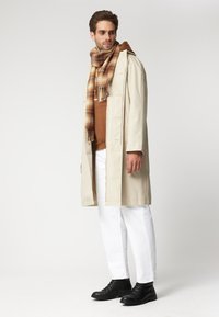 Beżowy trencz, brązowa bluza z kapturem, białe dżinsy i czarne botki na kostkę. Szalik ma wzór w kratkę z frędzlami.