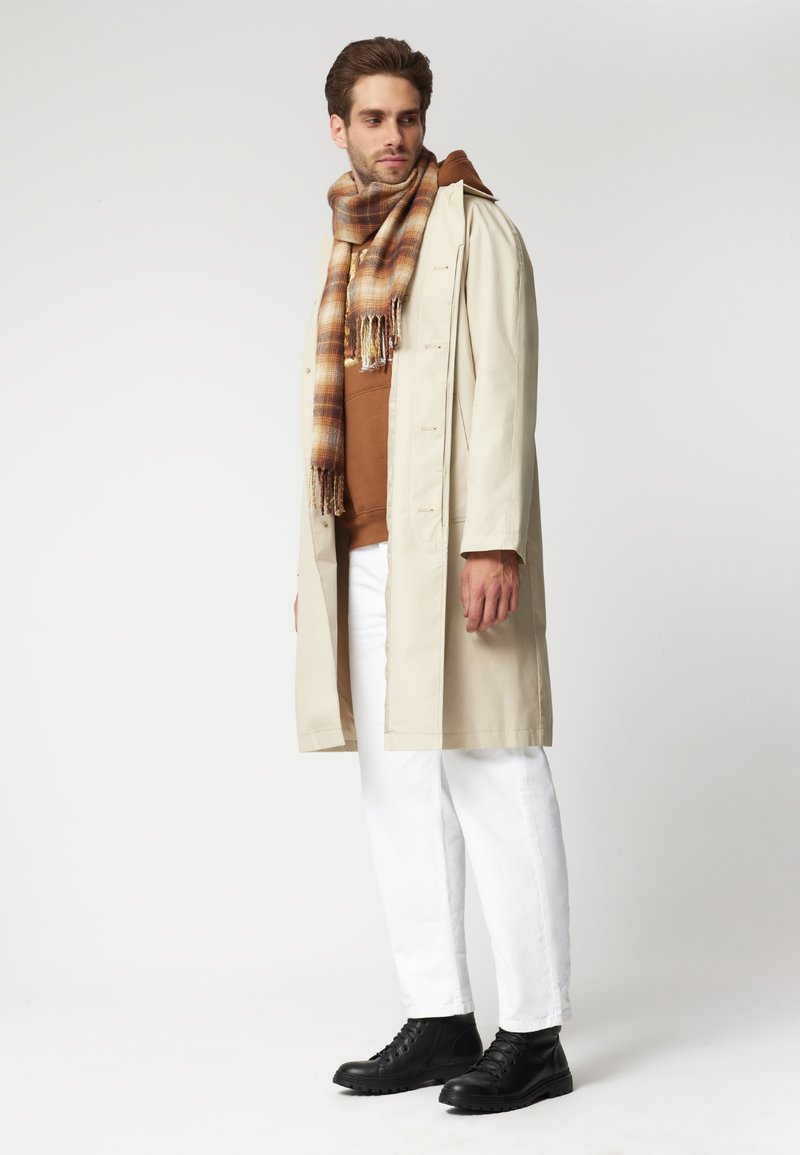 Beżowy trencz, brązowa bluza z kapturem, białe dżinsy i czarne botki na kostkę. Szalik ma wzór w kratkę z frędzlami.