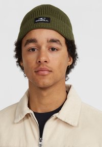 Beanie a coste verde oliva con un patch nero con il logo, indossato da una persona in giacca in corda crema sopra un top scuro.