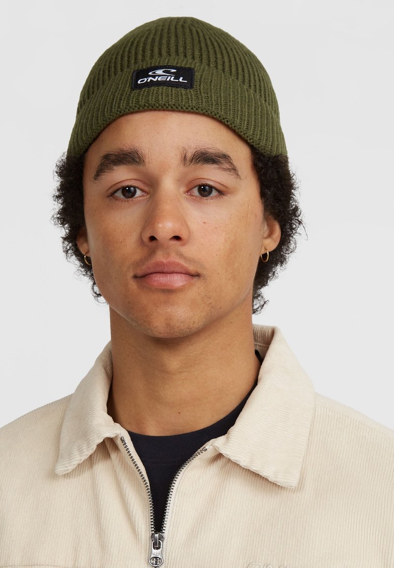Beanie a coste verde oliva con un patch nero con il logo, indossato da una persona in giacca in corda crema sopra un top scuro.
