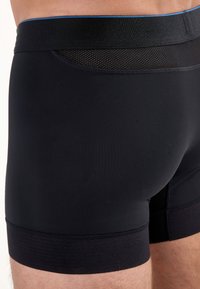 Schwarze Boxer Shorts aus glattem Stoff, mit einem kontrastierenden elastischen Bund und einem Netzpaneel für Atmungsaktivität. Ohne Muster oder Akzente.