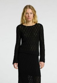 Top lungo nero a maniche lunghe realizzato in pizzo testurizzato con motivo a rombi, caratterizzato da un collo rotondo e un orlo a strati.