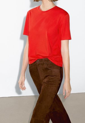 Personne portant un t-shirt rouge vif à manches courtes rentré dans un pantalon en daim marron taille haute, debout contre un mur blanc uni.